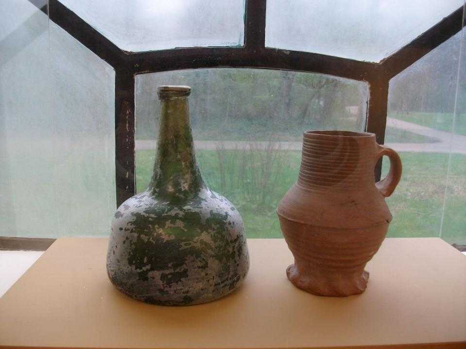 Foto van twee flessen op een foto: links de groen glazen wijnfles, 1875-1925 en rechts de steengoed drinkkan uit Siegberg uit Duitsland uit 1300-1325. Beiden gevonden op het voormalig land van Beinsdorp